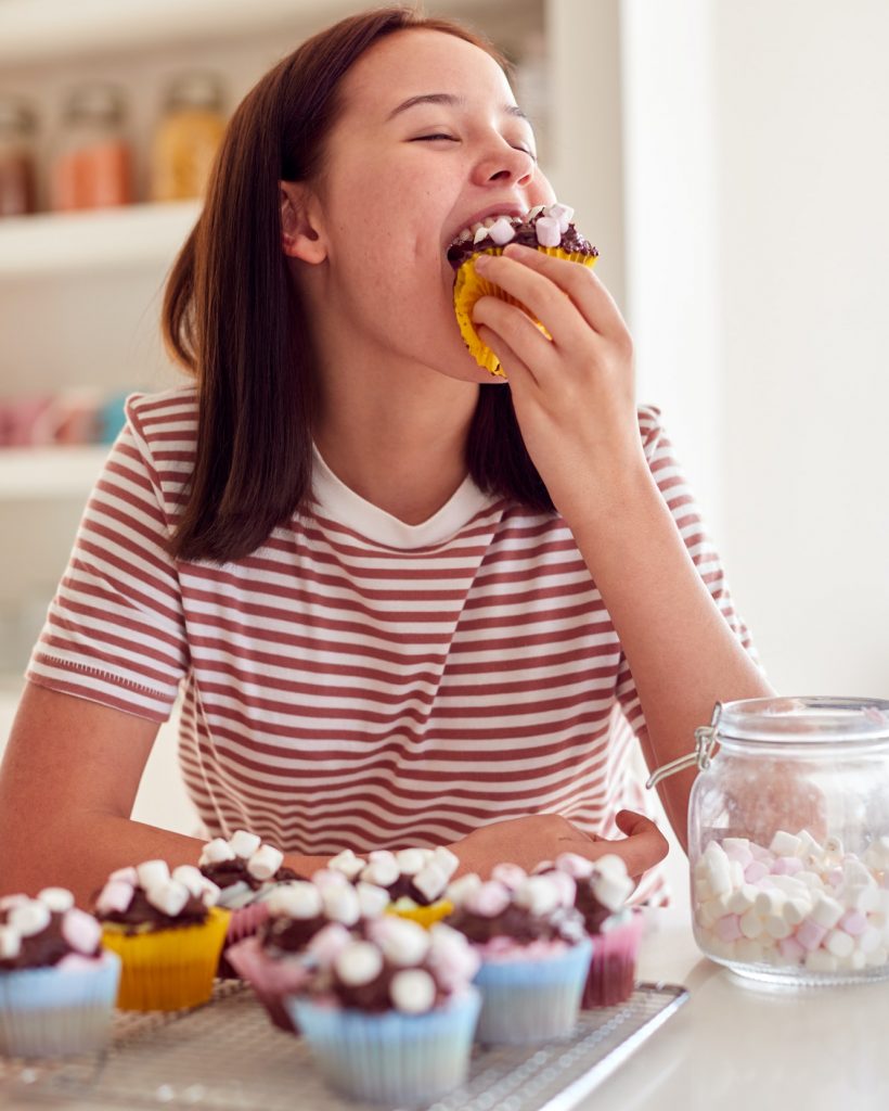 Teenage Girl Enjoying Eating Chocolate Cupcakes In Kitchen At Home